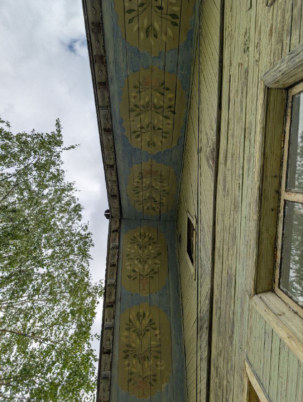 Вид вдоль свеса крыши с повторяющимися круглыми растительными медальонами и потёртой зелёной краской на фоне неба и кроны берёзы