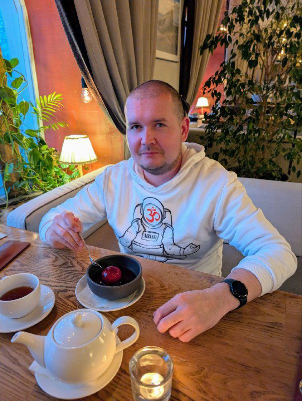 Гость в зале держит десерт с ярким красным шариком — можжевеловым мороженым; на столе видны чашки чая и свеча, уютная сервировка.