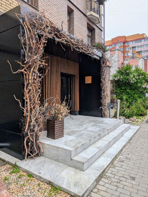 Вход в ресторан с тёмной деревянной отделкой и лианами, ступеньки и городской жилой дом на заднем плане — фасад Roomi в Архангельске.