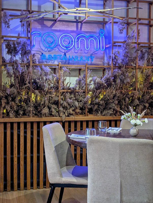 Интерьер ресторана с неоновой вывеской Roomi, сервированным столом и декоративной зеленью на стене — атмосфера заведения внутри.