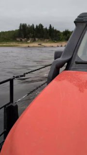 Кадр с близкого расстояния: боковая часть красного автомобиля и цепь, крепящая палубу парома, на фоне реки и дальнего берега с лесом.