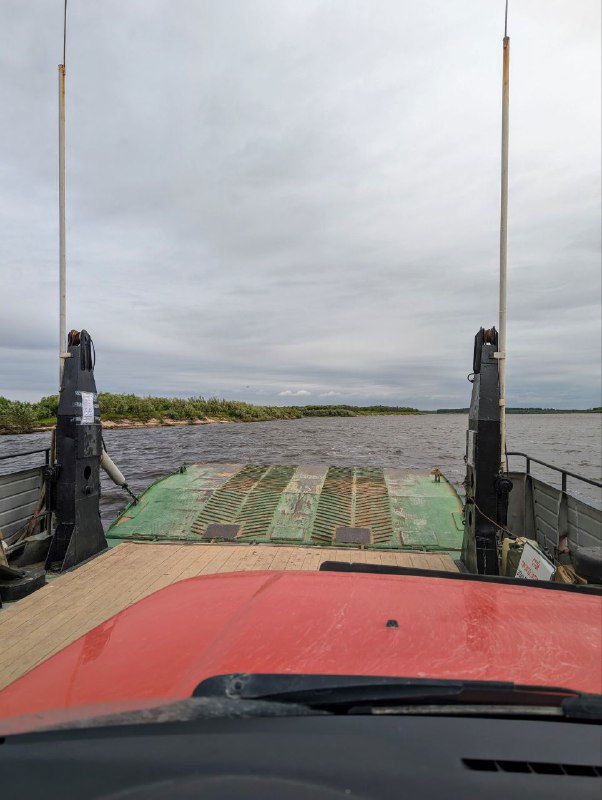 Вид с палубы парома на въездную рампу и настил, по которому подъезжает автомобиль; оттенки неба и реки под пасмурной погодой.