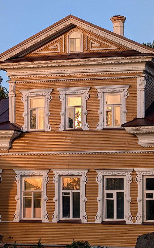 Фрагмент фасада деревянного дома в Вологде с декорированными наличниками и орнаментом, отражающимся в тёплом закатном свете на стеклах