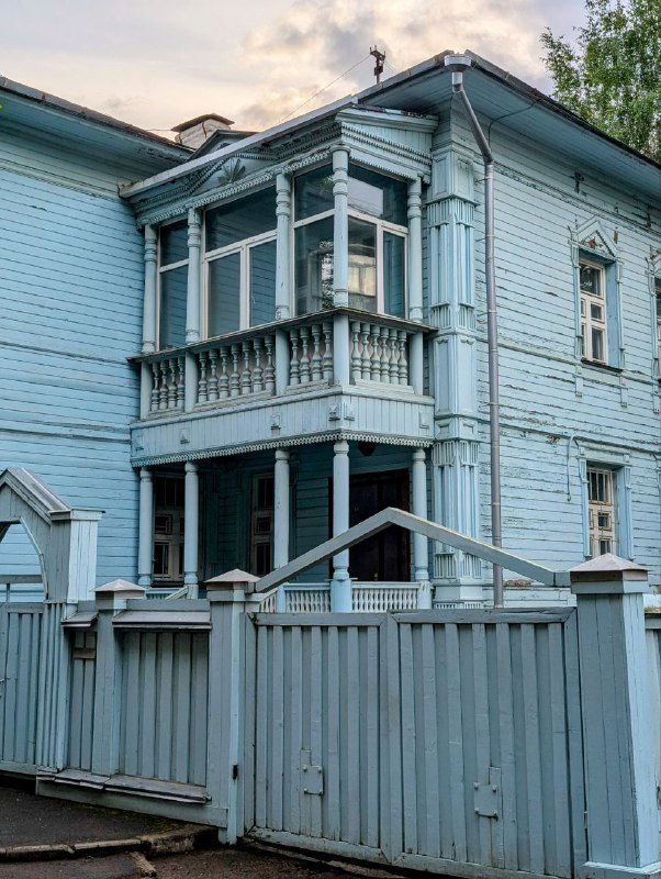 Голубой деревянный дом в Вологде с характерным закрытым балконом и резными стойками, ограждение и ворота того же тона на переднем плане