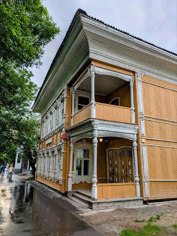 Жёлто-коричневый двухэтажный деревянный дом в Вологде с резными наличниками и веранной частью, угол здания и дождливая мокрая тротуарная плитка