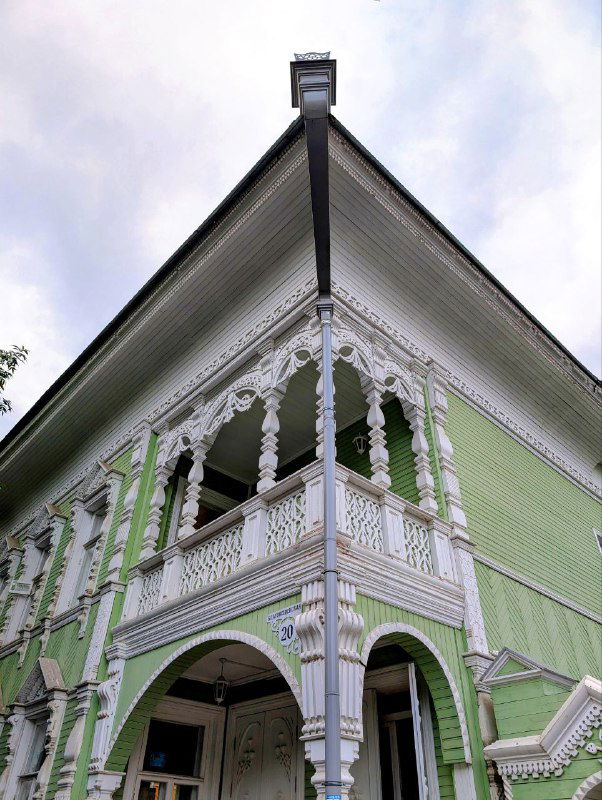 Бледно-зелёный деревянный дом с богатой ажурной резьбой на балконе и фронтоне, вид снизу подчёркивает сложный резной декор и глубину проёмов