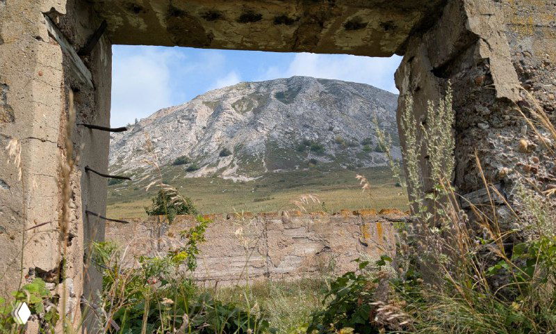 Вид на шихан через проём разрушенного здания ГУЛАГа, в кадре бетонные руины и известняковая гора на фоне лёгких облаков.