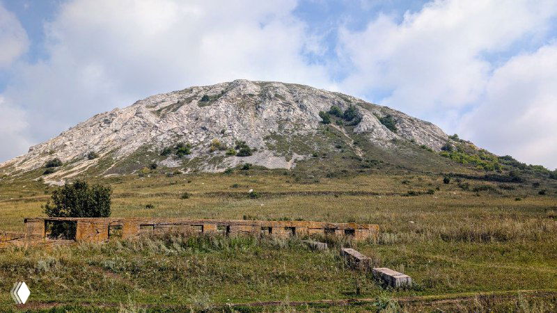 Широкий план шихана с руинами и остатками строений у подножия, ровная трава и дневное небо; и следы прежней промышленной деятельности.