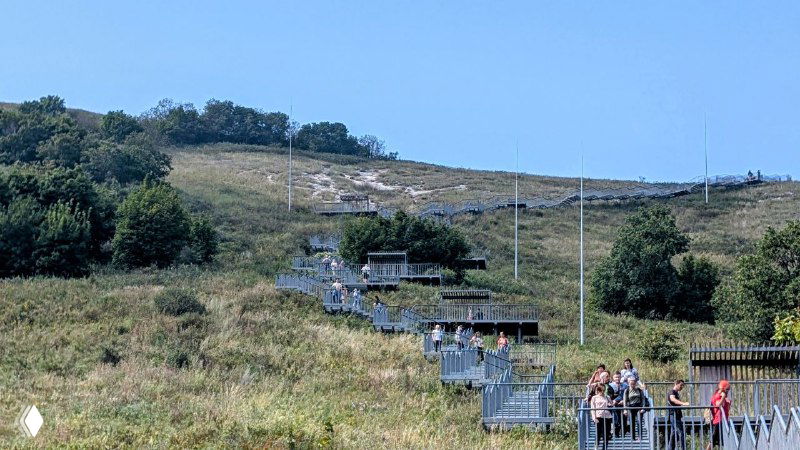 Лестница и ограждения ведут к вершине шихана, туристы спускаются и поднимаются, вокруг полевые травы и ясное небо.