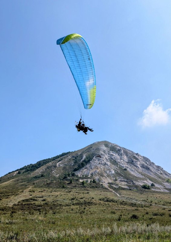 Мотопараплан в полёте над шиханом на фоне голубого неба; парапланерист и купол парашюта контрастируют с горой внизу.