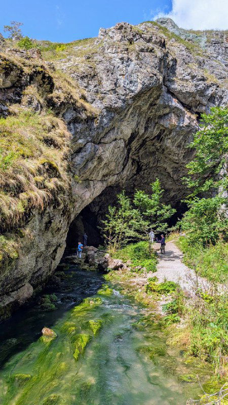 Вход в пещеру Шульган‑Таш: скальный свод и речка у подножия, летний ландшафт с тропой, с которой начинается путь к инфоцентру и дальше к пещере.