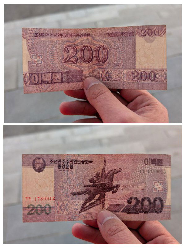 Две стороны купюры номиналом 200 вон, показанные рукой на фоне — пример корейских банкнот, которые туристам нельзя вывозить из КНДР