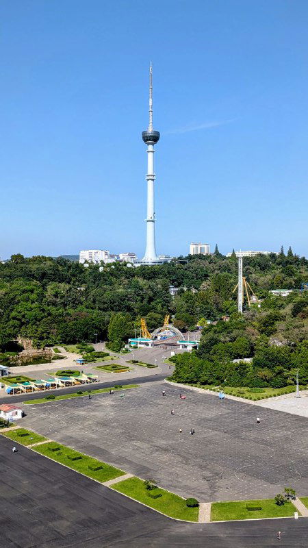 Телевизионная башня Пхеньяна вдалеке: панорамный вид с площади у арки, зелёные насаждения и свободная территория перед башней.