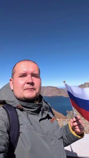 Селфи у озера Чон: турист держит флаг на фоне синей воды и скалистых хребтов кратера Пэкту под ясным небом.