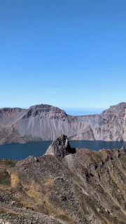 Скала и утёс на переднем плане, за ними ровная поверхность озера Чон и окружение кратера Пэкту под голубым небом.