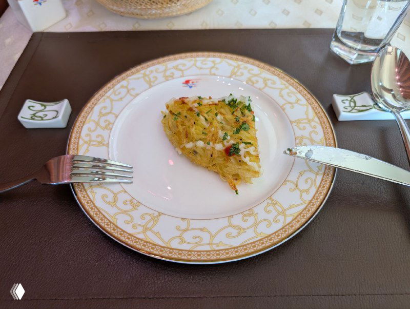 Небольшой кусок картофельной запеканки на фарфоровой тарелке с золотым узором и столовыми приборами — ресторанная подача.