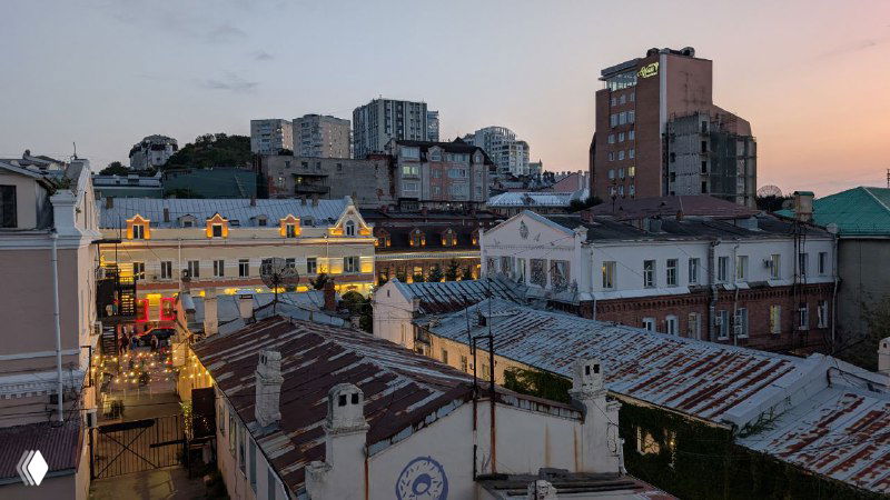 Закатный вид на крыши и освещённые улицы Владивостока: смесь старой застройки, холмов и вечернего света, панорамный городской мотив.