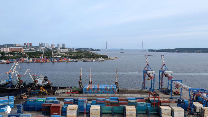 Вид на портовые контейнеры и крановую технику на фоне пролива и удалённого вантового моста в дальнем плане Владивостока.