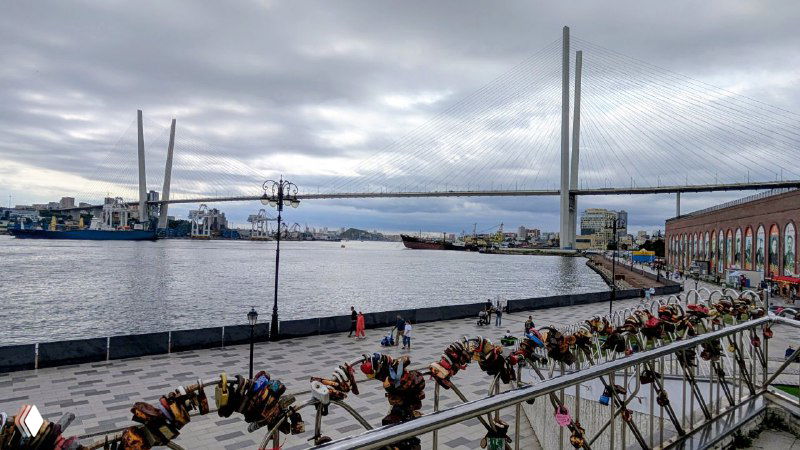 Морская набережная с видом на опоры вантового моста, люди и ограждения вдоль причала на фоне пролива в ясный день.