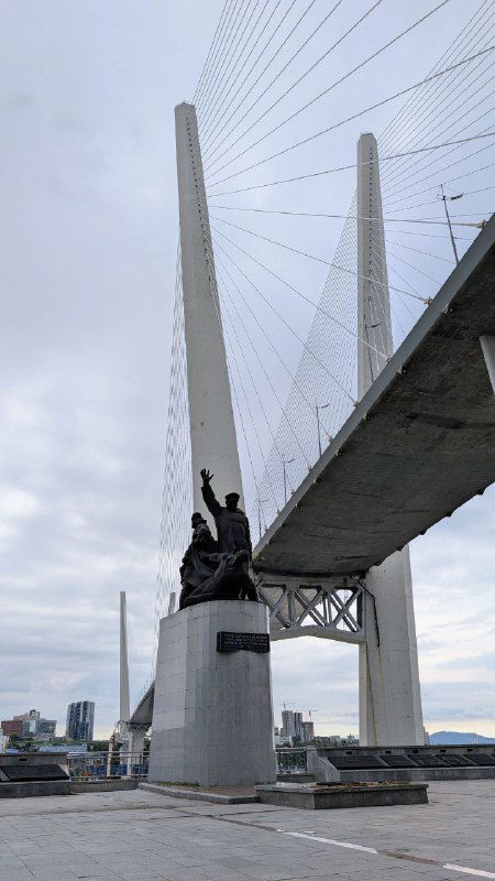Крупный план пилонов Золотого моста и памятника Морякам торгового флота у основания, вид снизу на архитектуру и стальные тросы.