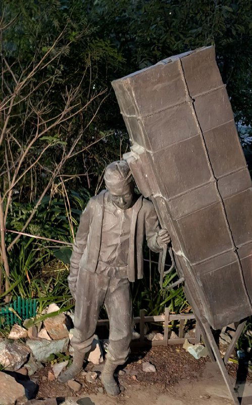 Скульптура рабочего (кула) с тяжёлым грузом во дворе Миллионки на фоне зелёных кустов — художественная инсталляция района.