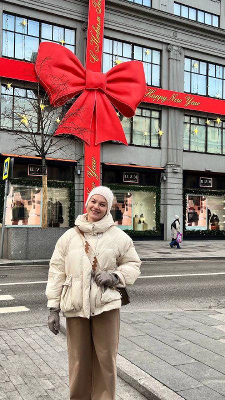 Женщина в тёплой одежде стоит на пешеходной улице перед огромным красным бантом — городской витринный декор и прогулочная сцена зимнего центра.