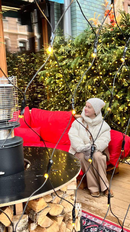 Уличное кафе с красным диваном и гирляндами: женщина в тёплой куртке сидит у декоративного уличного обогревателя, сцена городского уюта зимой.