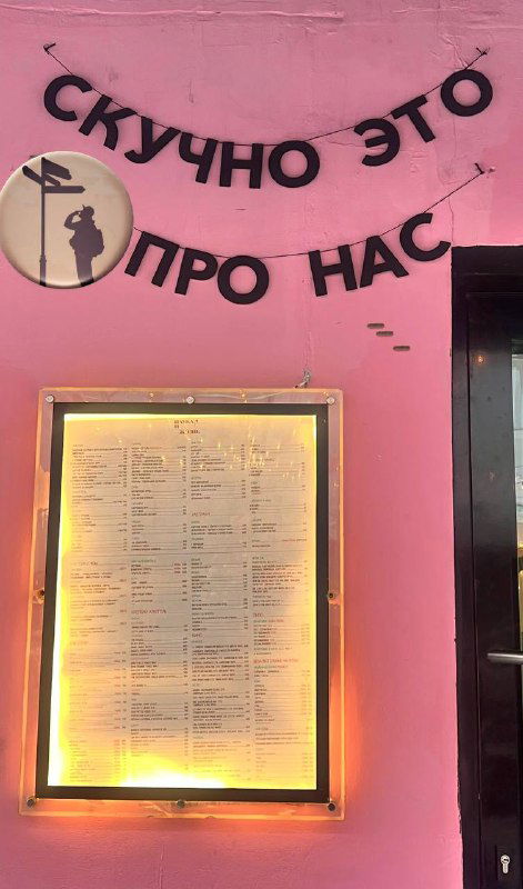 Розовая стена с надписью «Скучно это про нас» и подсвеченной рамкой меню — ироничная вывеска и атмосферный городской кадр с дизайном фасада.