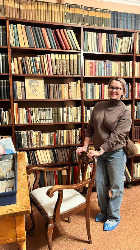 Полка с книгами в библиотеке для детей при доме Чуковского, деревянные стеллажи, кресло и посетительница на фоне книжной экспозиции и иллюстраций.