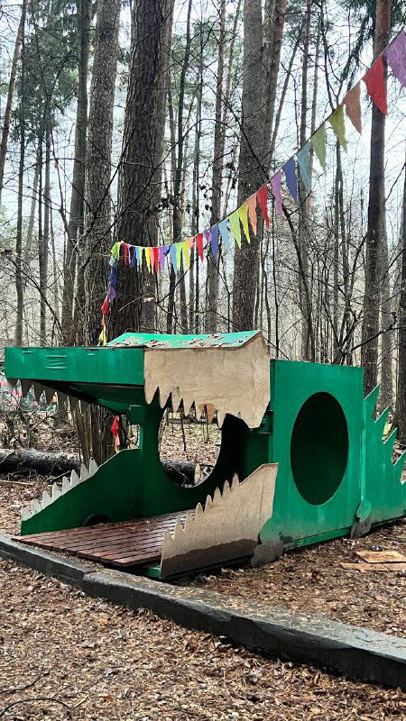 Большая деревянная игровая скульптура в виде крокодила на детской площадке перед домом Чуковского, окрашенная и украшенная флажками и играми для детей.