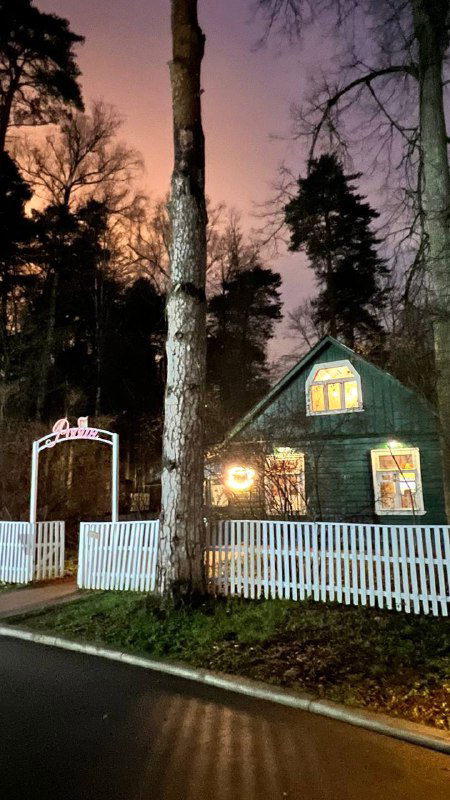 Небольшой деревянный дом с белым забором и подсветкой вечером — дом Чуковского в Переделкино на фоне высоких деревьев и сумеречного неба.