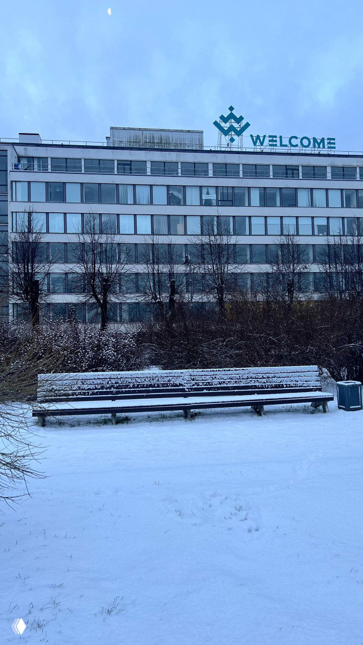 Снежная скамья на фоне многоэтажного корпуса с вывеской «WELCOME», зимний городской парк с белым покровом на траве.