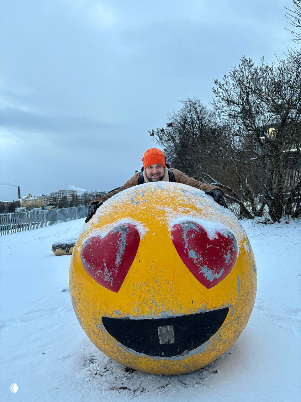 Яркая жёлтая скульптура-смайлик с глазами в форме сердец, покрытая снежной пылью; рядом человек в оранжевой шапке улыбается.