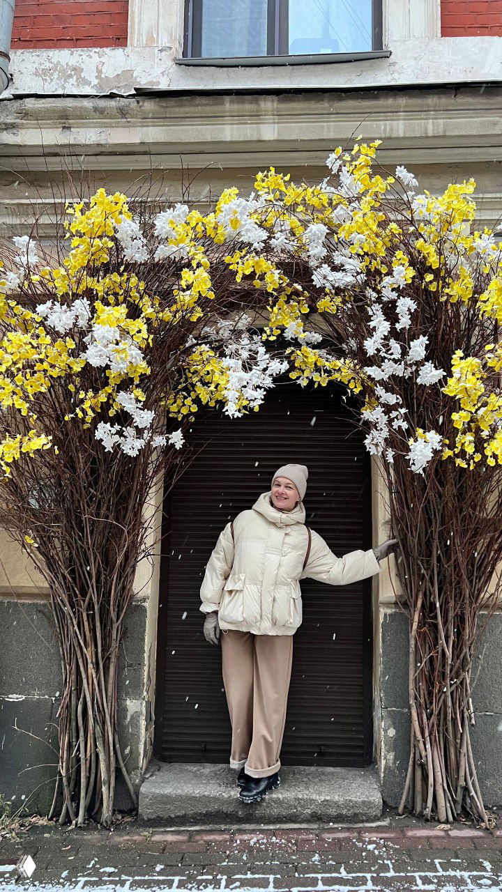 Человек в светлой куртке позирует на фоне снежной площади и круглой башни, на снегу видны следы и простор вокруг.