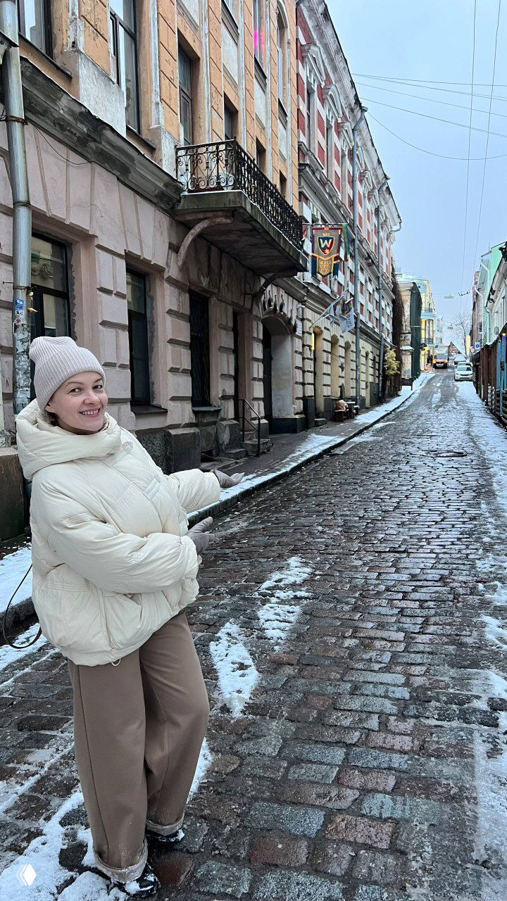 Уютная улочка с булыжной мостовой, заснеженные фасады и улыбающийся человек в шапке, указывающий на дорогу вперёд.