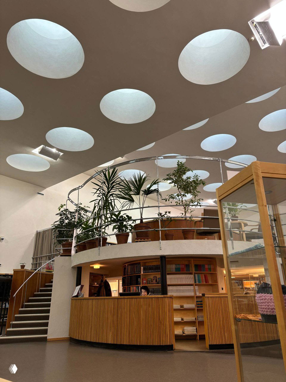 Внутренний вид читального зала библиотеки Аалто с круглыми световыми люками в потолке, растениями на галерее и ресепшн‑стойкой у входа.