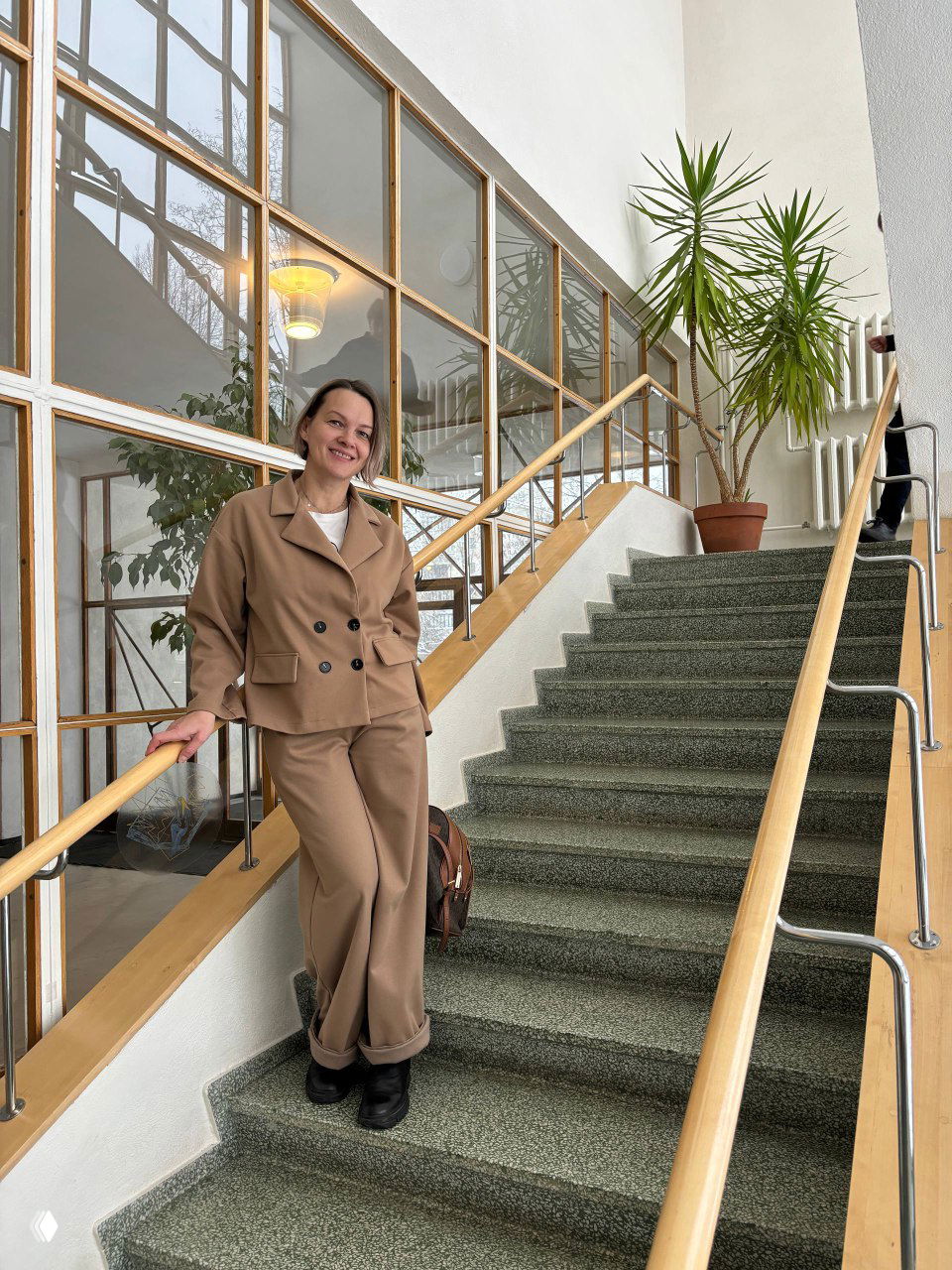 Посетительница в бежевом костюме на лестнице библиотеки Аалто; крупные окна дают вид на заснеженный парк с берёзами и создают спокойную атмосферу.
