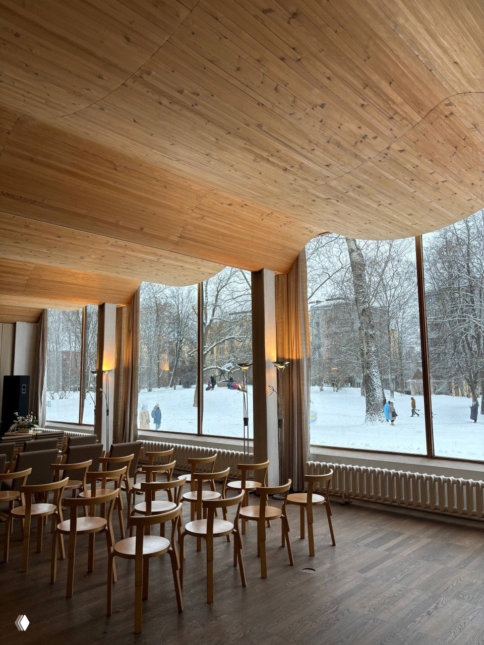 Концертный или актовый зал с тёплым деревянным потолком, рядами стульев и большими окнами на заснеженный городской пейзаж — пример продуманной общественной зоны.