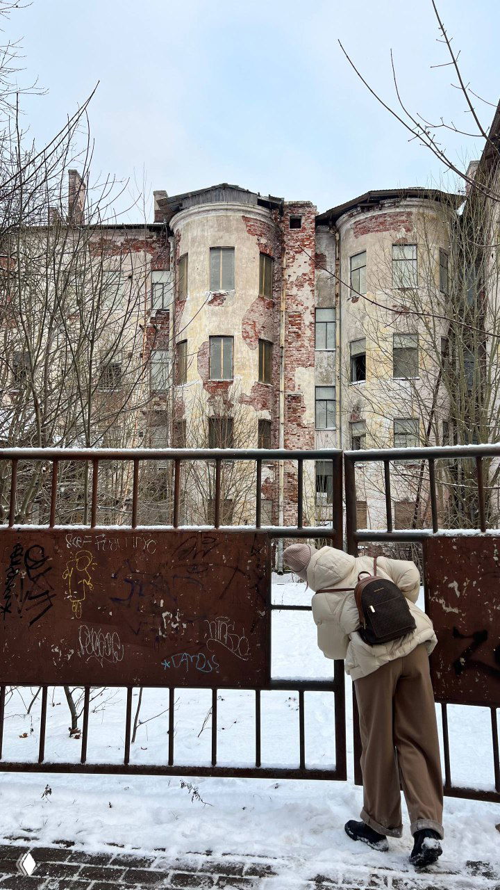 Величественный многоквартирный дом с облупившейся штукатуркой и кирпичными вставками, вид с забора и фигура прохожего, заглядывающего во двор.