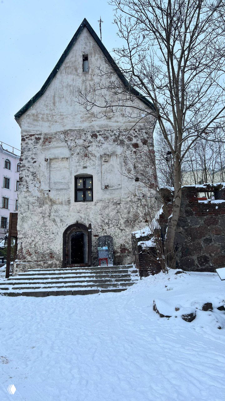 Старинное каменное здание с крутой зубчатой крышей и простыми окошками, заснеженные ступени и вход в низком зимнем свете.