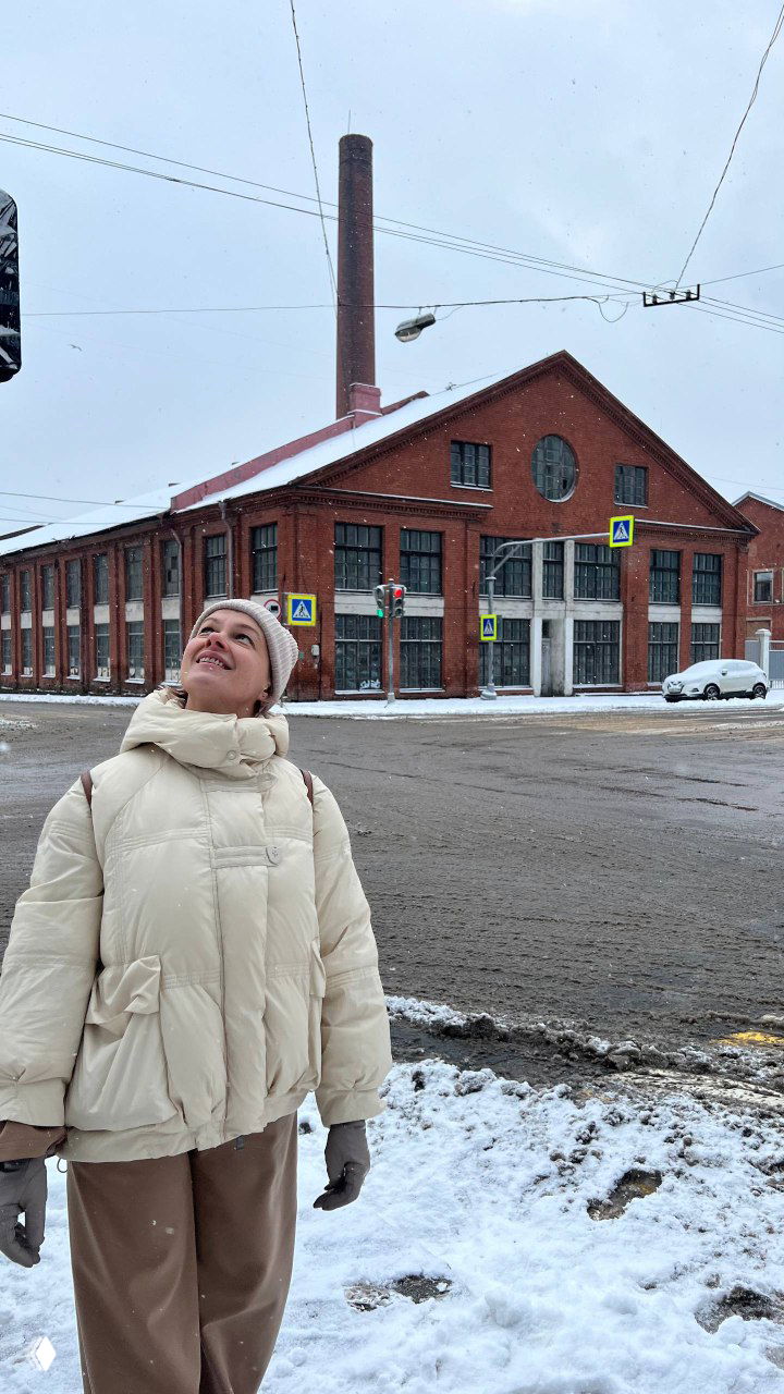 Красный кирпичный индустриальный корпус с высокой трубой на фоне пасмурного неба; на переднем плане улыбающаяся женщина смотрит вверх.