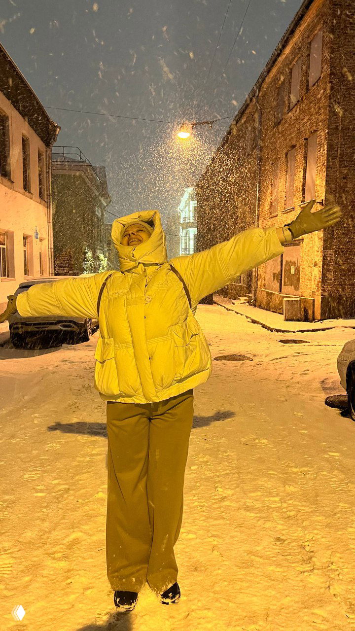 Ночная улица, сильный снегопад и женщина в светлой куртке с поднятыми руками, тёплые уличные фонари создают золотистый свет в кадре.