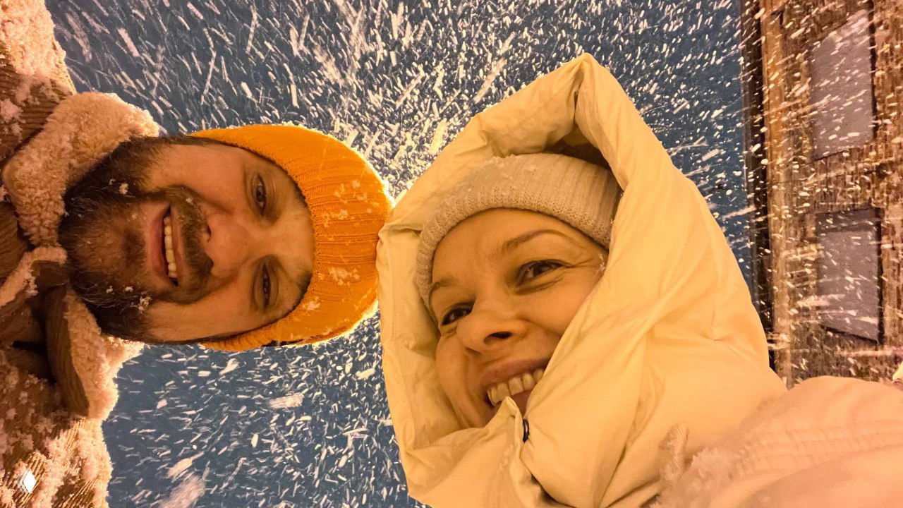 Крупный селфи‑кадр двух людей в зимней одежде под сильным снежным потоком, улыбающиеся лица и ёмкая атмосфера прогулки.