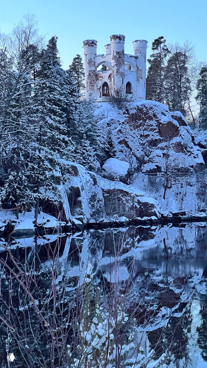 Старое романтическое строение на высокой скале в Монрепо, окружённое соснами и снегом, отражается в почти спокойной воде.