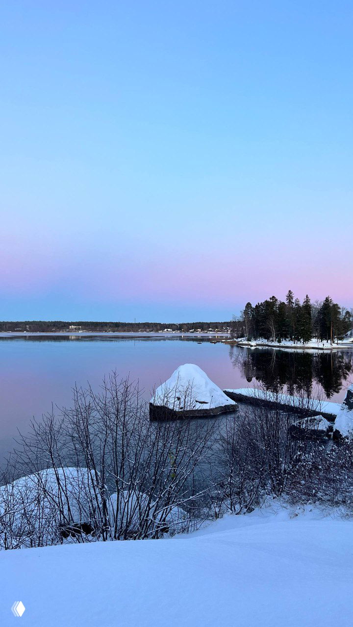 Береговая линия залива с большим заснеженным камнем на переднем плане и мягким розово‑голубым небом — тихий зимний рассвет.