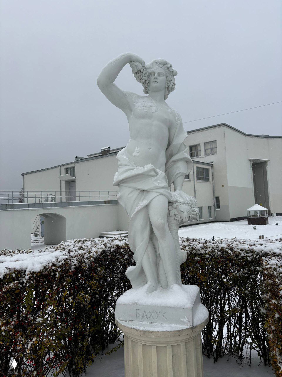 Белая скульптура Бахуса на постаменте перед зданием музея Монрепо; вокруг падает снег, у подножия видны ступени и посетитель.