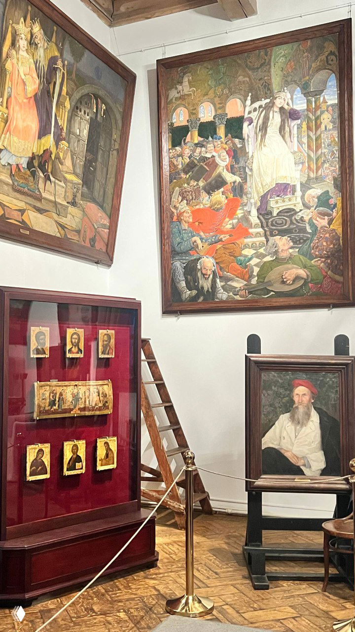 Угол экспозиции: большие полотна на стенах, иконы в витрине и предметы быта на фоне деревянной лестницы и потолочных балок.