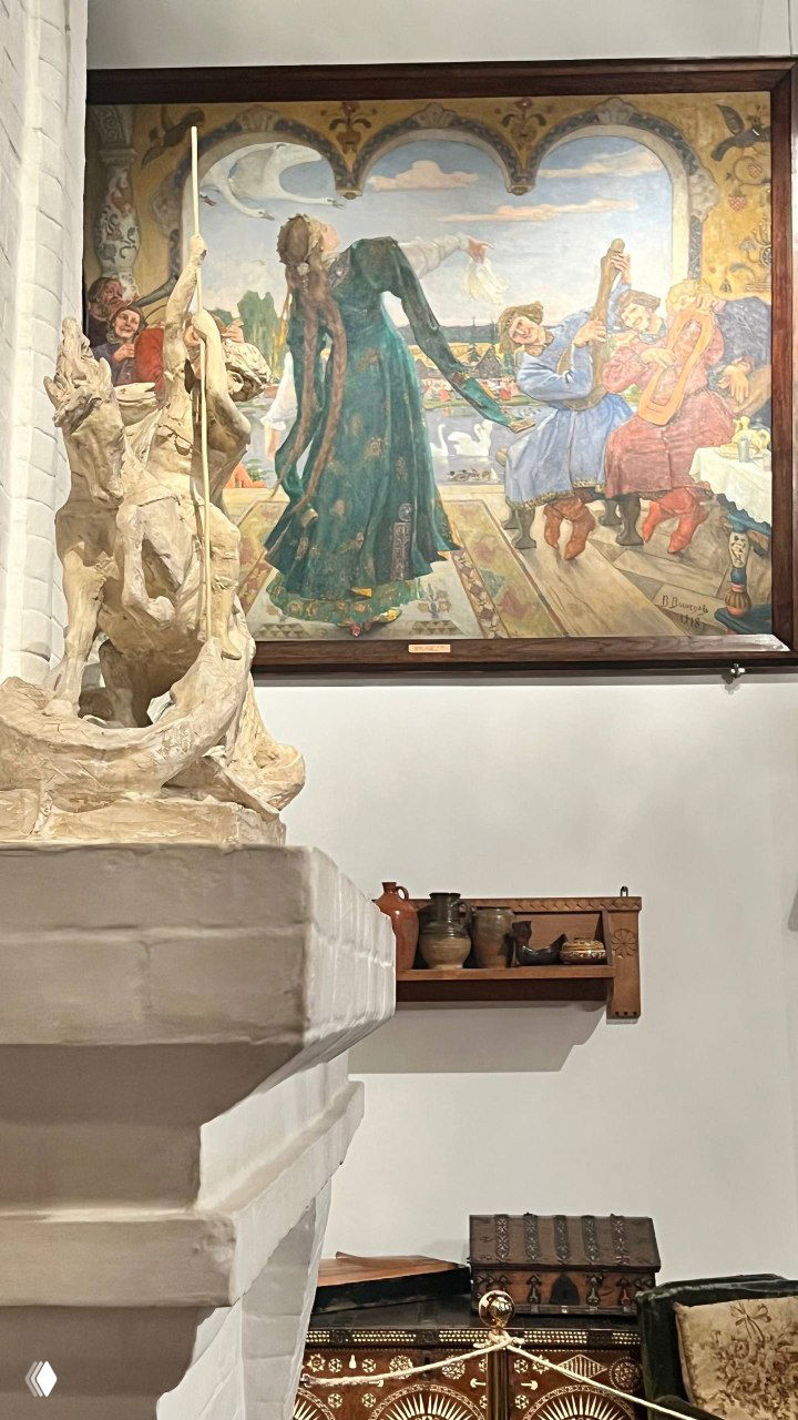 Скульптурный фрагмент на переднем плане и живописная картина на стене позади; интерьер в неорусском духе музея Васнецова.