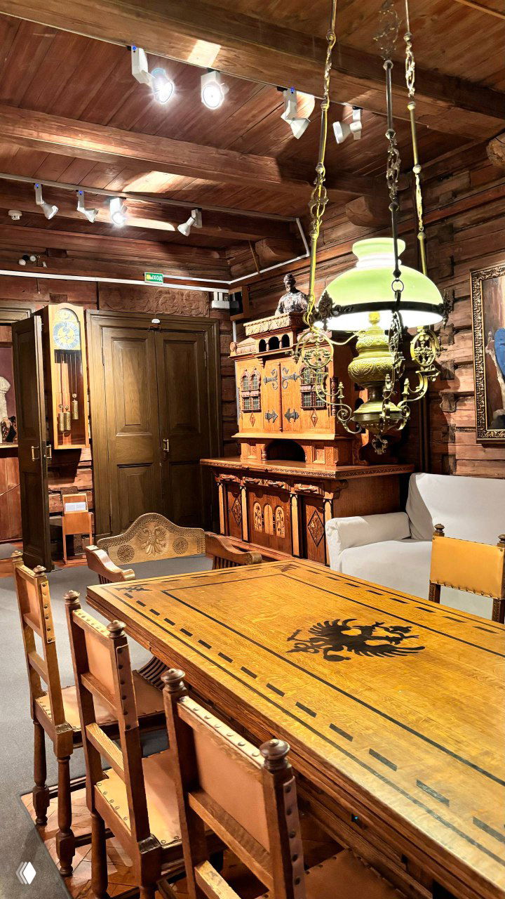 Интерьер комнаты с деревянной мебелью и резными элементами, длинный стол и лампы — пример домашней обстановки в музее‑усадьбе.