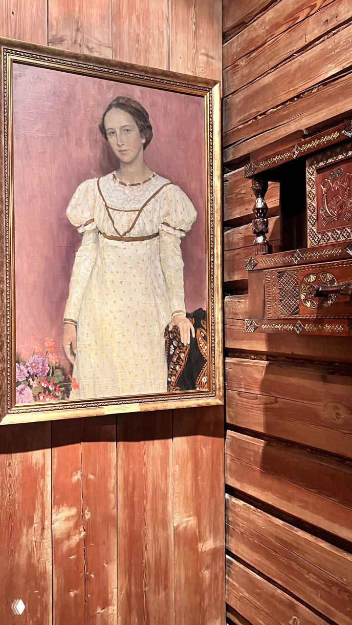 Портрет молодой женщины в золотой раме на фоне деревянной панели стены музея; экспозиция подчёркивает бытовой контекст.